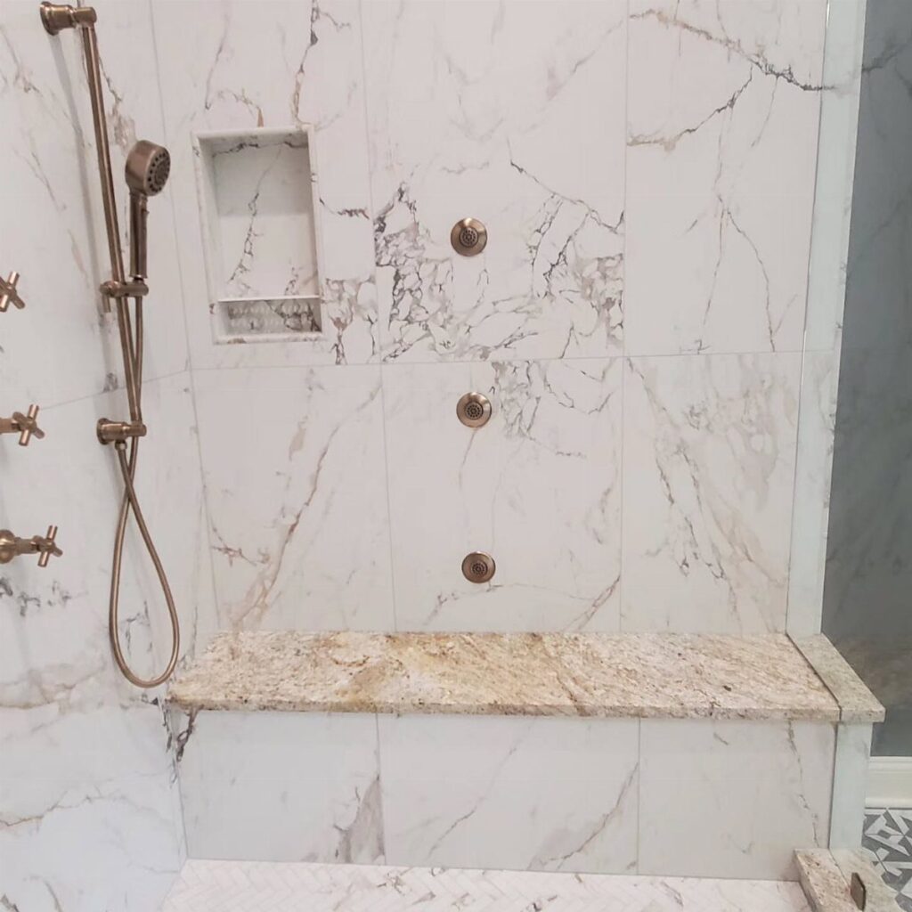 Modern walk-in shower with white marble walls, gold fixtures, and a granite bench.