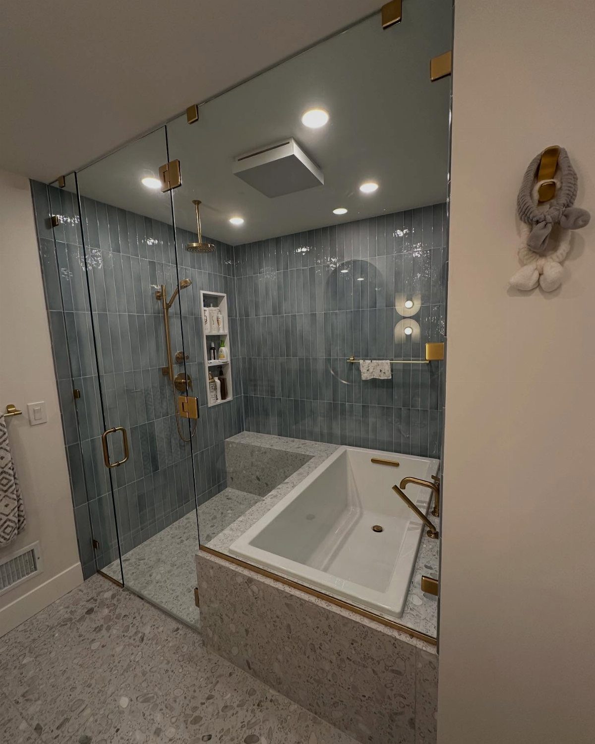 Modern bathroom with blue-grey tiled walls, glass shower enclosure, and white soaking tub.