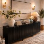 Black fluted console table with a white vase and lamp against a paneled wall.
