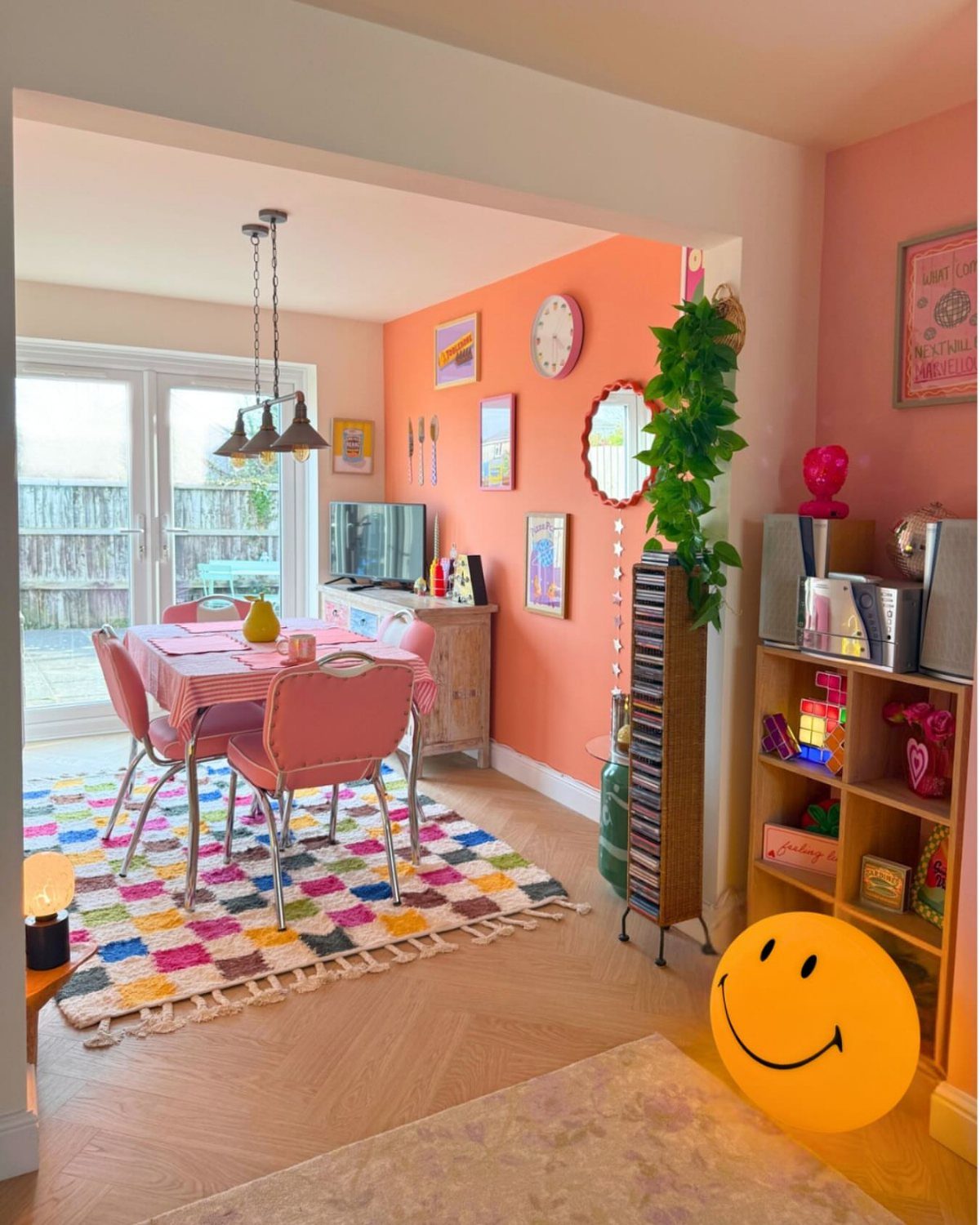 Colorful dining room with pink furniture, orange walls, and a large smiley face lamp.