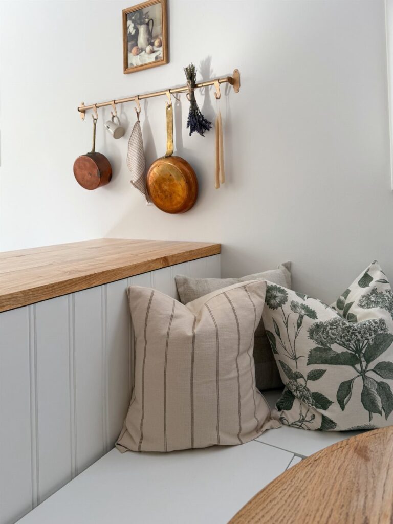 Kitchen nook with white bench, decorative pillows, and copper pots on a gold rail.
